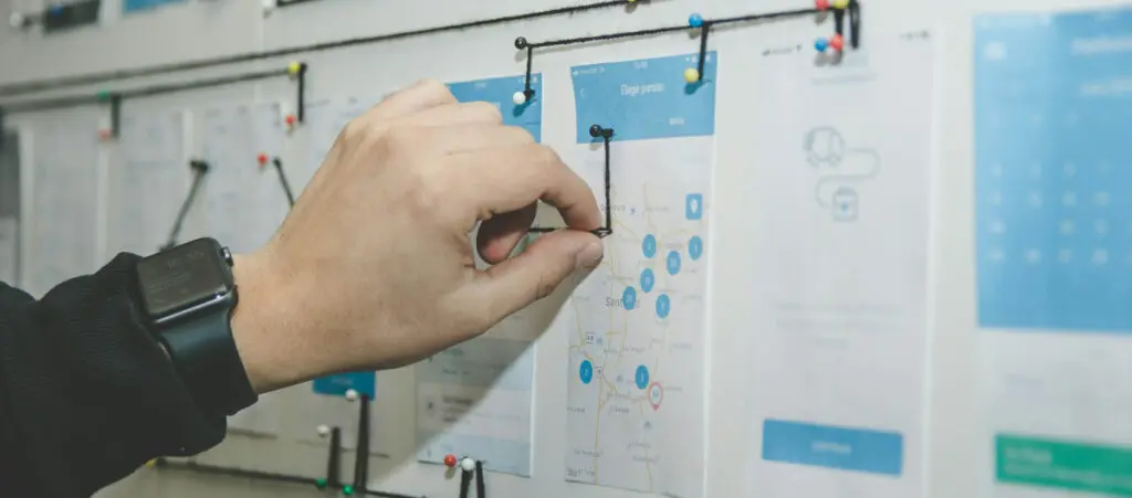 a closeup of a hand placing pins with strings on a board with various printed pieces of paper detailing a plan