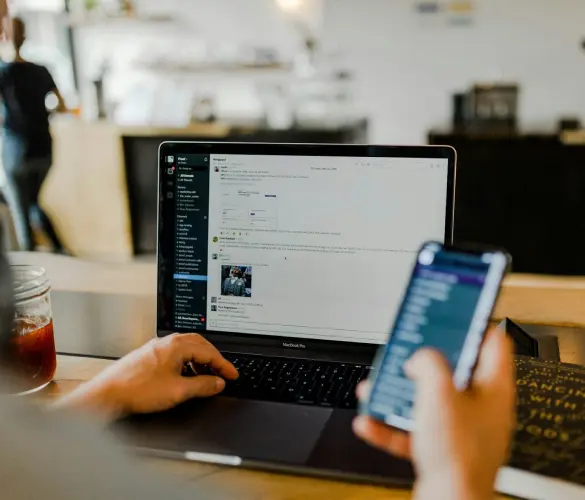 an office worker sits on a chair and holds a smartphone while using the Slack app on his computer with the other hand