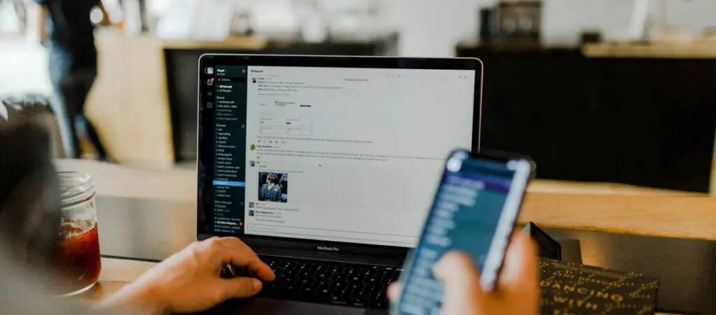 an office worker sits on a chair and holds a smartphone while using the Slack app on his computer with the other hand