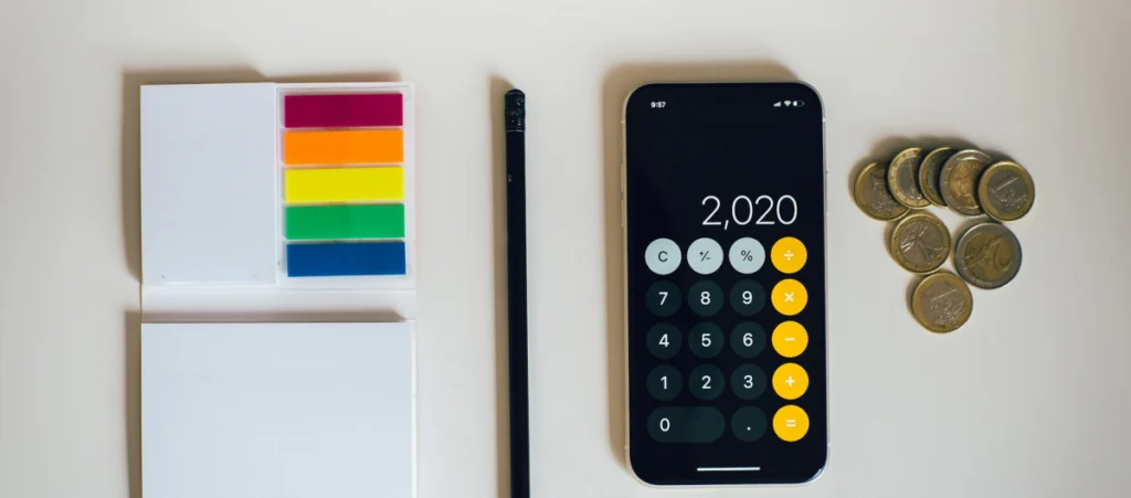 an overhead view of various tools, including coins, a smartphone displaying a calculator, a pen, and paper